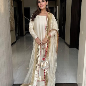May include: A cream-coloured, long-sleeved dress with gold and red trim, paired with a matching shawl and small purse. The outfit features intricate gold detailing and is worn with gold bangles. The ensemble is suitable for special occasions.