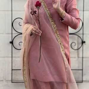 May include: A dusty rose-colored outfit, including a long-sleeved top and matching skirt, is accented with gold trim and a sheer scarf. A red rose is held in the hand. The outfit has a pearl necklace and gold bracelets.