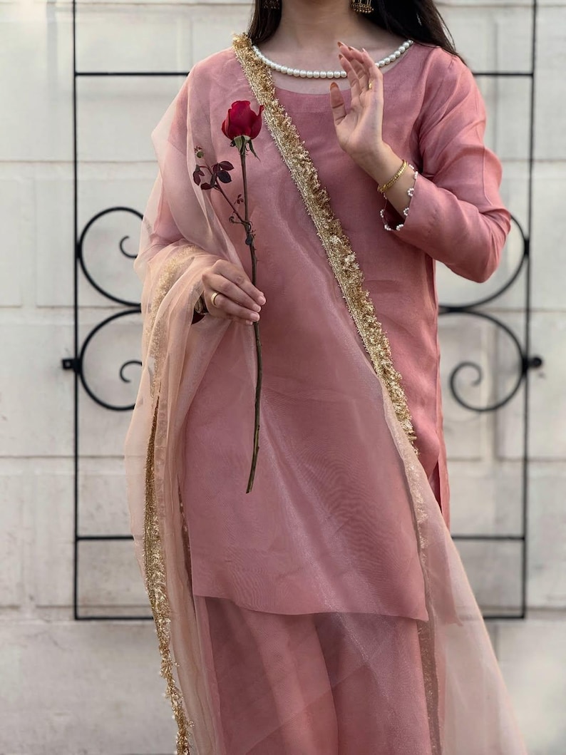 May include: A dusty rose-colored outfit, including a long-sleeved top and matching skirt, is accented with gold trim and a sheer scarf. A red rose is held in the hand. The outfit has a pearl necklace and gold bracelets.