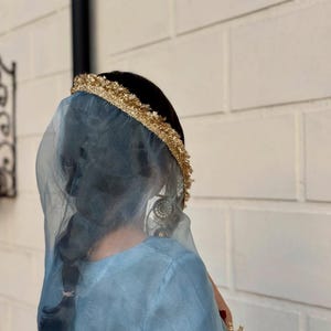 May include: A person wearing a light blue dress and veil, with a gold-colored headpiece. The veil is sheer, and the headpiece is adorned with gold trim. The person has dark hair styled in a braid. The background is a white brick wall.