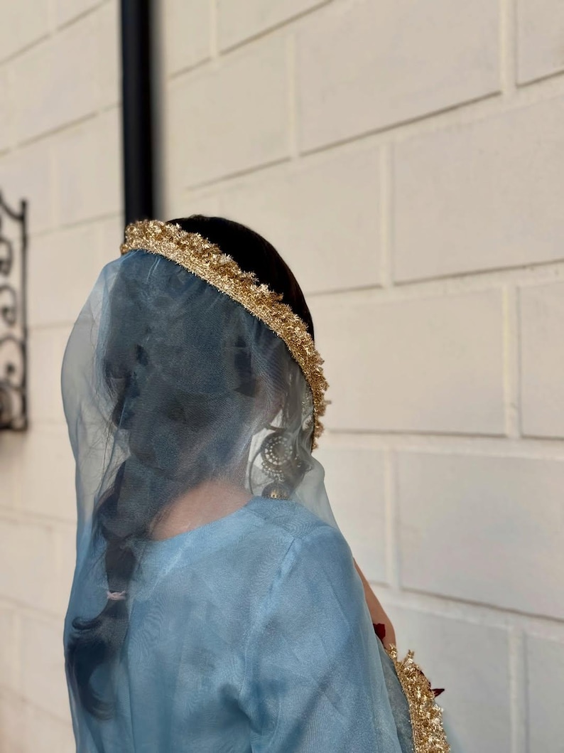 May include: A person wearing a light blue dress and veil, with a gold-colored headpiece. The veil is sheer, and the headpiece is adorned with gold trim. The person has dark hair styled in a braid. The background is a white brick wall.