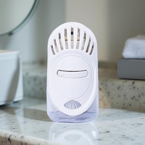 May include: A white air freshener with a clear liquid reservoir. The device has a dial with settings from 1 to 3 and a slotted top. It is designed to release fragrance into the air. The product is sitting on a countertop.