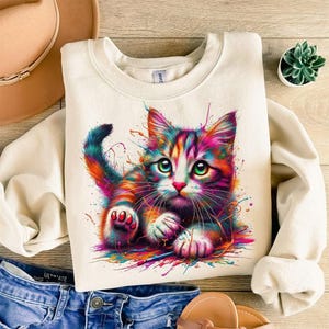 May include: Cream-colored sweatshirt featuring a colorful, artistic illustration of a cat. The cat has vibrant rainbow-colored fur and green eyes, with paint splatter effects around it. The sweatshirt is paired with blue denim jeans and a brown hat.