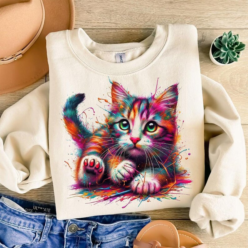 May include: Cream-colored sweatshirt featuring a colorful, artistic illustration of a cat. The cat has vibrant rainbow-colored fur and green eyes, with paint splatter effects around it. The sweatshirt is paired with blue denim jeans and a brown hat.