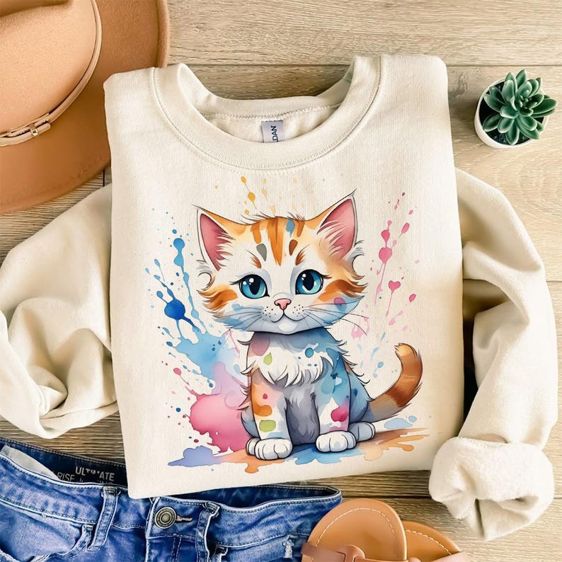 May include: Cream-colored sweatshirt featuring a watercolor illustration of a cute cat with orange and white fur and blue eyes. The cat is surrounded by colorful paint splatters in blue, pink, and purple. A casual, comfortable style.