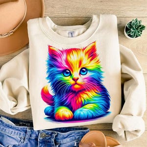May include: Cream-colored sweatshirt featuring a vibrant, rainbow-colored kitten graphic. The kitten has large blue eyes and fluffy fur in shades of red, orange, yellow, green, blue, and purple. The sweatshirt is made of a soft material.