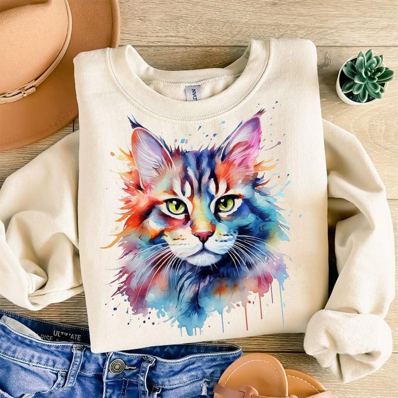 May include: Cream-colored sweatshirt featuring a watercolor painting of a cat's face. The cat's fur is a mix of blue, red, orange, and purple, with green eyes. The design has a splash paint effect. The sweatshirt is folded on a wooden surface.