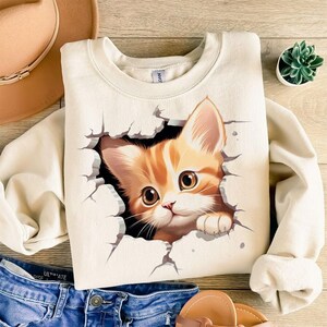 May include: Cream-colored sweatshirt featuring a graphic of a ginger kitten breaking through a wall. The kitten has large brown eyes and is looking forward. The sweatshirt is paired with blue jeans and a hat.