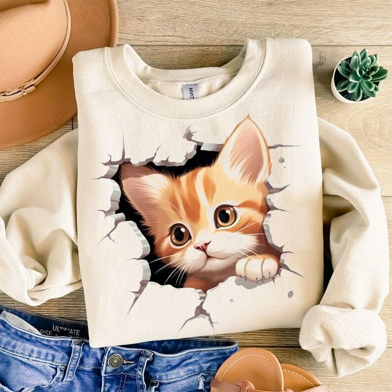 May include: Cream-colored sweatshirt featuring a graphic of a ginger kitten breaking through a wall. The kitten has large brown eyes and is looking forward. The sweatshirt is paired with blue jeans and a hat.