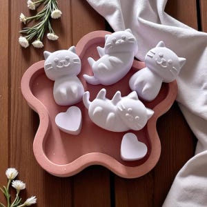 Pode incluir: Uma bandeja rosa em forma de flor contém quatro estatuetas de gatos brancos em várias poses e dois objetos em forma de coração. Os gatos têm traços faciais simples. A bandeja está sobre uma superfície de madeira, com tecido branco e flores ao fundo.