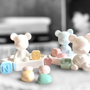 Op de afbeelding: Een verzameling pastelkleurige berenbeeldjes en letterblokken. De beren zijn wit, lichtblauw en roze. De blokken spellen letters en hebben harten en andere vormen. Het algemene thema is babydecoratie.