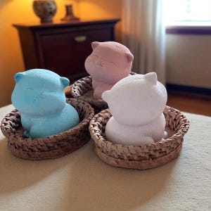 May include: Three cat ornaments in blue, pink, and white, each in a small woven basket. The cats have a smiling expression. The round baskets are brown, and the ornaments sit on a neutral surface.