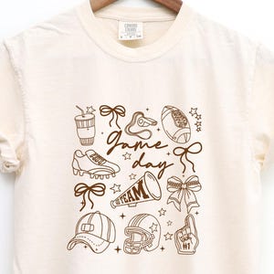 May include: Cream-colored t-shirt with a brown graphic design featuring football-related illustrations and the words "Game Day" and "Team". The design includes a football, helmet, cleats, and other sports-themed elements.