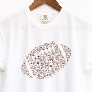 May include: White t-shirt with a brown floral football design. The football is made up of small flowers and leaves, creating a unique and artistic look. The shirt is hanging on a wooden hanger.