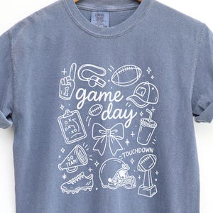 May include: A blue short-sleeved t-shirt with a white graphic design featuring football-related icons and the words "game day." The design includes a football, helmet, and other sports-themed elements. The shirt is hanging on a wooden hanger.