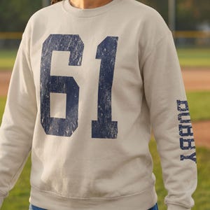 May include: A beige crewneck sweatshirt featuring a large, distressed navy blue number 61 printed on the front. The left sleeve displays the word BUBBY in a matching distressed navy blue font, oriented vertically along the forearm area.