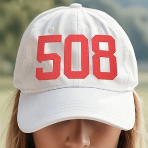 May include: A white baseball cap featuring the number "508" in red block letters. The cap has a curved peak and a button on the crown. The background is blurred, showing green foliage.