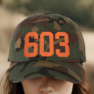 Peut inclure: Casquette de baseball camouflage avec le numéro "603" en lettres orange. La casquette présente un design classique à six panneaux avec une visière incurvée. L'arrière-plan est une scène extérieure floue.