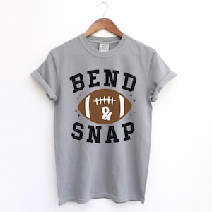 May include: A gray t-shirt with the words "BEND & SNAP" in black block letters. A brown football graphic with white laces and the "&" symbol is centered between the words. The shirt is hanging on a wooden hanger.