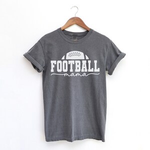 May include: A gray t-shirt with rolled-up sleeves hangs on a wooden hanger. The shirt features a white graphic of a football above the word "FOOTBALL" and the word "mama" in a script font.