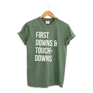 Könnte beinhalten: Olivgrünes T-Shirt mit dem weißen Aufdruck "FIRST DOWNS & TOUCHDOWNS". Das T-Shirt hängt an einem Holzbügel vor weißem Hintergrund. Die Ärmel sind hochgekrempelt.