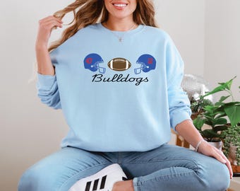 Könnte beinhalten: Hellblaues Sweatshirt mit Football-Motiv. Das Design zeigt zwei blaue Helme mit den Nummern 47 und 12 sowie einen braunen Football. Das Wort "Bulldogs" ist in einer Schriftart unter den Grafiken geschrieben.