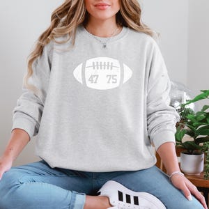 May include: A light gray sweatshirt with a white football graphic. The football graphic has the numbers 47 and 75 inside. The sweatshirt is a casual style, suitable for everyday wear.