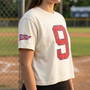 May include: A cream-coloured short-sleeve t-shirt featuring a large, distressed red number nine on the front with a blue outline. The right sleeve displays a small red pennant graphic with the word TIGERS printed in white letters.