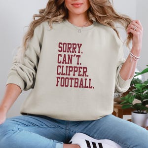 May include: A beige sweatshirt with the text "SORRY. CAN'T. CLIPPER. FOOTBALL." in maroon block letters. The sweatshirt is long-sleeved and has a crew neck. The person in the image is wearing blue jeans and white sneakers.