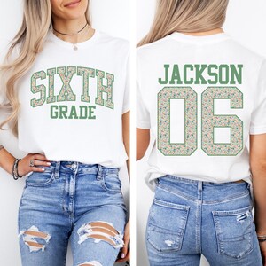 May include: White t-shirt with the words "SIXTH GRADE" in a floral design on the front. The back of the shirt displays "JACKSON 06" in a similar floral pattern. The shirt is a classic crew neck style.