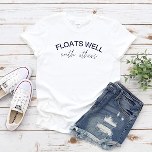 May include: A white t-shirt with the text "FLOATS WELL with others" in navy blue script, paired with denim shorts and white canvas sneakers. The items are arranged on a light wood-grain surface, with a small green plant.