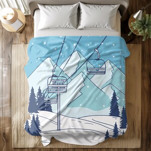 Può includere: Una coperta blu e bianca con un design di seggiovia, montagne e neve che cade. La coperta ha un tema invernale con una seggiovia, pini e un paesaggio innevato.