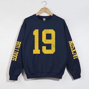 May include: A navy blue crewneck sweatshirt featuring the number 19 in a distressed yellow font on the chest. The word BULLDOGS is printed in yellow on the left sleeve, and the word JACKSON is printed in yellow on the right sleeve.