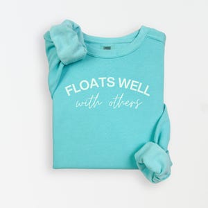 May include: A turquoise sweatshirt with the phrase "FLOATS WELL with others" printed in white. The sleeves are rolled up. The sweatshirt is made of a soft material.