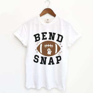 May include: White t-shirt with the words "BEND & SNAP" in black block letters above and below a brown football graphic. The shirt is hanging on a wooden hanger.