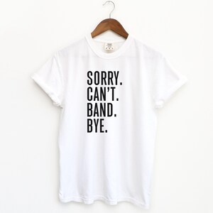 Può includere: T-shirt bianco con la scritta nera "SORRY. CAN'T. BAND. BYE.", appeso a una gruccia di legno. La maglietta è realizzata in un materiale morbido e ha un classico girocollo.