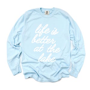 May include: A light blue long-sleeve shirt with the phrase "life is better at the lake" in white script. The shirt has a crew neck and long sleeves with ribbed cuffs. The shirt is made of a soft material.