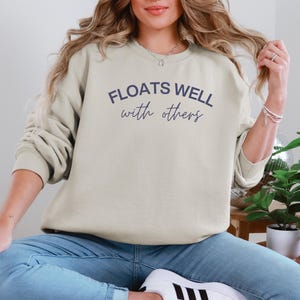 Sand-toned unisex crewneck that says “Floats Well With Others,” ideal for summer lounging and group float trips.