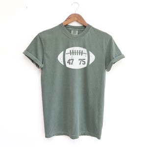 May include: A sage green t-shirt with a white football graphic. The football graphic has the numbers 47 and 75 inside. The shirt is hanging on a wooden hanger against a white background.