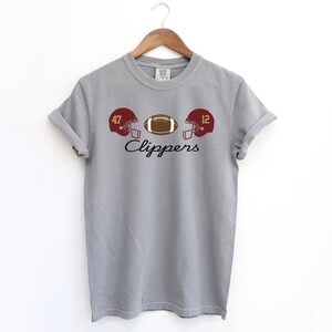 May include: A gray t-shirt with a football theme. The shirt features a brown football, two maroon football helmets with the numbers 47 and 12, and the word "Clippers" in a script font. The shirt is hanging on a wooden hanger.