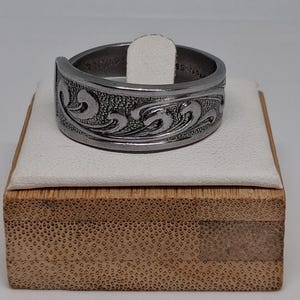 May include: A silver-toned metal ring featuring a continuous embossed scrollwork pattern along the outer band. The interior of the ring band displays visible engraved text, including the characters SS, and the ring has a slightly open design.