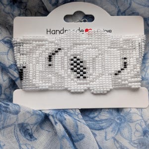 Peut inclure: Un ras-du-cou blanc perlé avec le mot "LOVE" épelé en perles noires. Le choker est présenté sur une carte blanche avec les mots "Handmade Love". Le fond est un tissu à motifs floraux bleus et blancs.