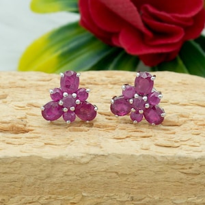 Könnte beinhalten: Ein Paar Ohrstecker mit Anhäufungen von ovalen, tiefrosa Edelsteinen. Die Ohrringe sind in Silber gefasst und in einem floralen Design angeordnet. Der Hintergrund umfasst eine rote Rose und grüne Blätter.