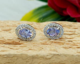May include: A pair of oval-shaped stud earrings. Each earring features a light purple gemstone surrounded by a halo of small, clear stones. The earrings are set in a silver-colored metal and rest on a textured, light brown surface.