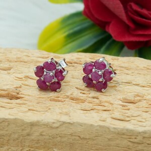 May include: A pair of silver stud earrings, each featuring a cluster of oval-cut, deep red gemstones arranged in a flower shape. The earrings are set against a light brown wooden surface, with a blurred background of a red rose and green leaves.