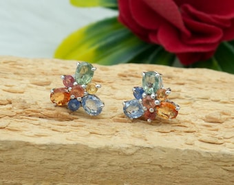 May include: A pair of stud earrings featuring a cluster of colorful gemstones. The earrings showcase a mix of oval and round-cut stones in shades of blue, orange, green, and yellow, set in a silver metal. The earrings are displayed on a wooden surface.