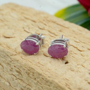 May include: A pair of silver stud earrings, each featuring a vibrant, oval-cut ruby gemstone. The rubies display a rich, deep pink hue, set in a classic, minimalist design. The earrings are presented on a light brown wooden surface.