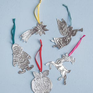 Puede incluir: Conjunto de adornos navideños plateados con cintas de colores. Los adornos incluyen una estrella fugaz, un ángel tocando la trompeta, un Papá Noel, una granada y un reno. Sobre un fondo azul claro.