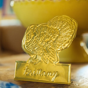 May include: A gold-colored metal place card holder shaped like a turkey with a fanned tail. The base features the name Brittany embossed in a cursive script font. The object has a reflective, metallic finish and stands upright on a flat surface.
