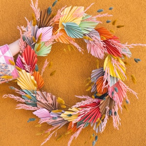 May include: A colorful paper wreath with a variety of colors, including pink, yellow, orange, and green. The wreath is made of paper fans and is decorated with dried flowers and grasses.